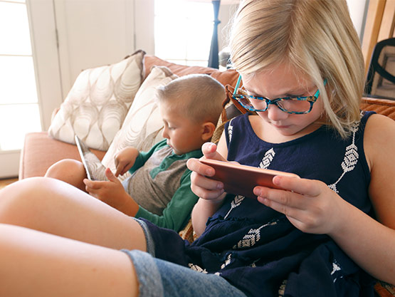 A little boy and girl sitting on a couch wearing glasses and looking at a phone and tablet